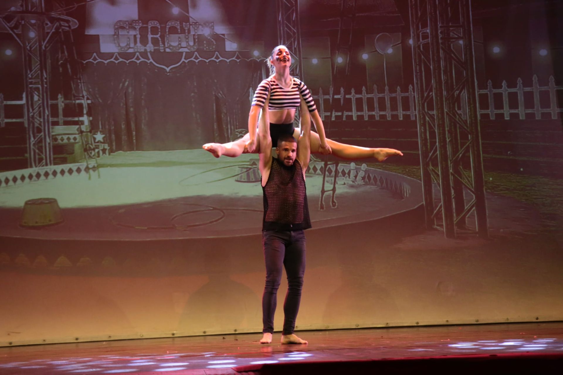 Su un palco con uno sfondo a tema circense, una persona solleva un'altra persona in posizione di spaccata sopra la sua testa.