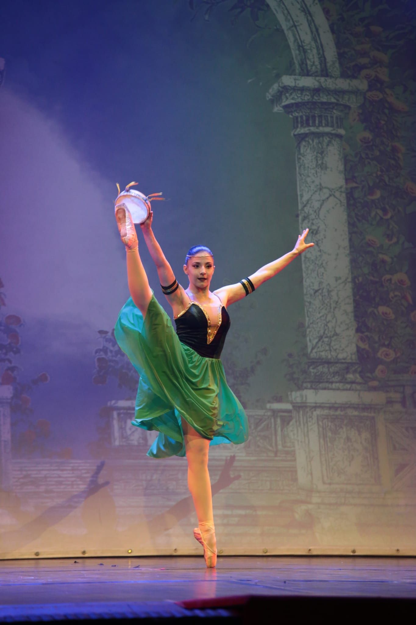 Una ballerina con gonna verde e corpetto nero si bilancia su una gamba mentre tiene in alto un tamburello sul palcoscenico di un teatro.