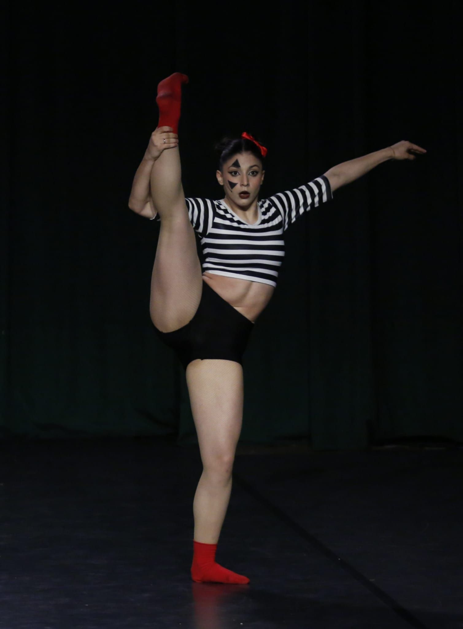 Una ballerina con una maglietta a righe e pantaloncini neri esegue un'estensione verticale della gamba sul palco, contro uno sfondo scuro.
