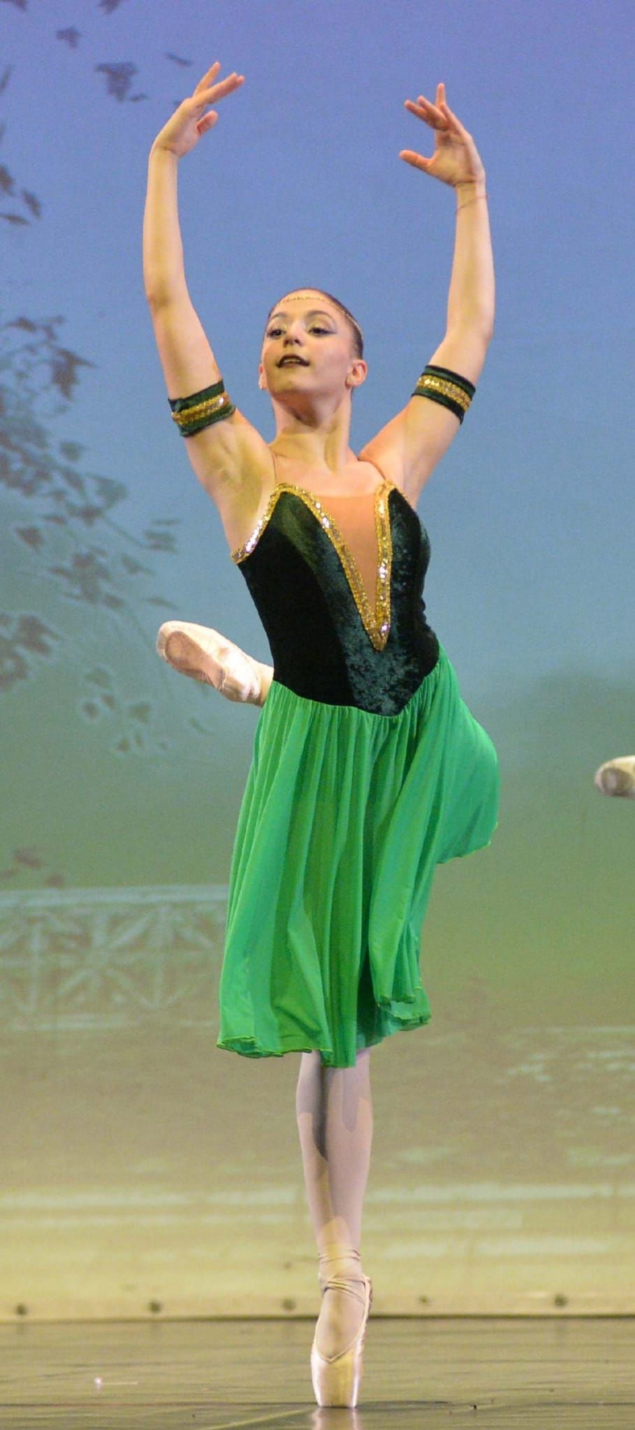 Una ballerina in costume nero e verde si esibisce sul palco con le braccia alzate, in equilibrio su una sola punta.