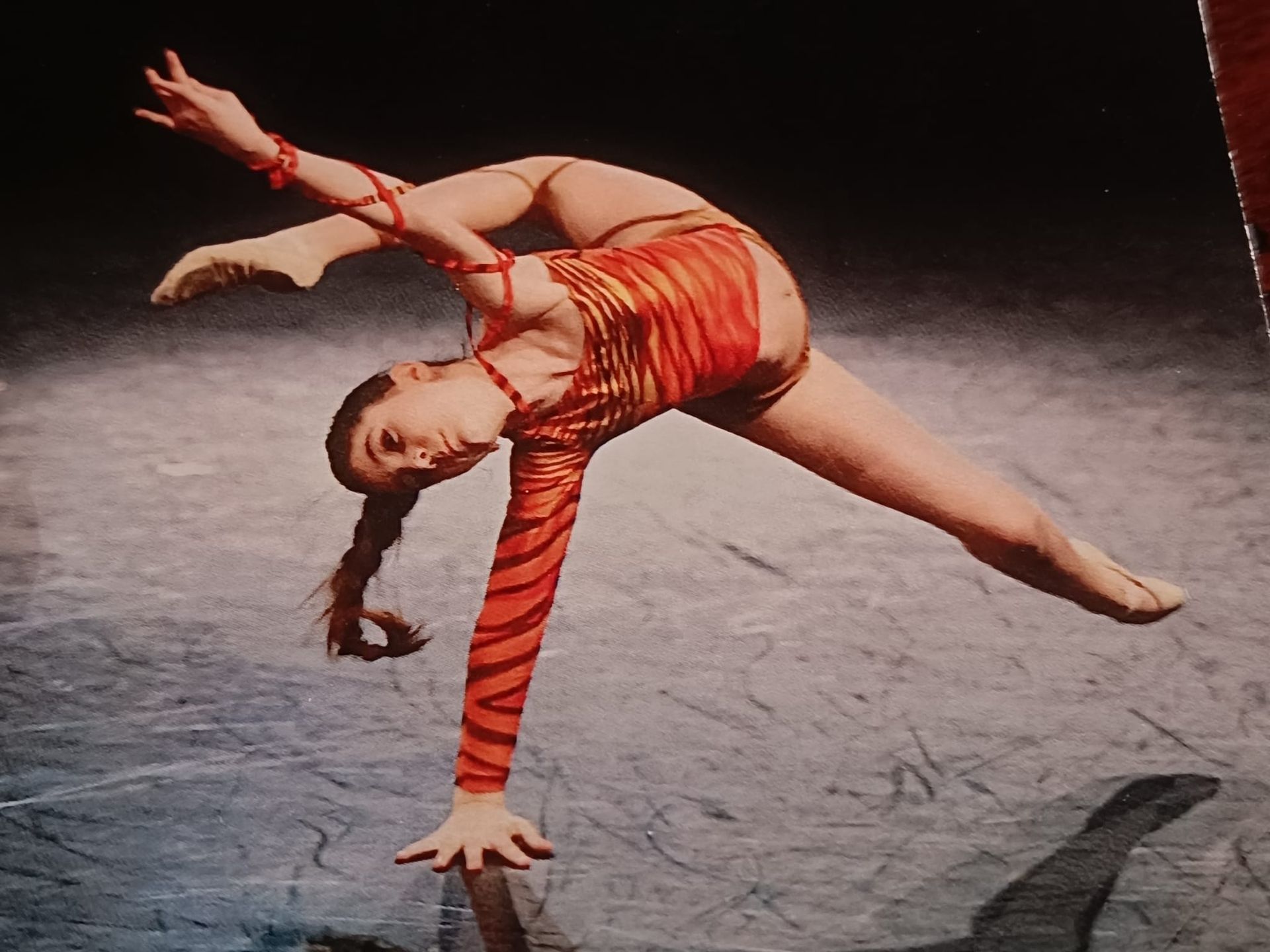 Una ballerina con un abito a righe arancioni esegue una posa flessibile con una sola mano su un palcoscenico scuro e riflettente.
