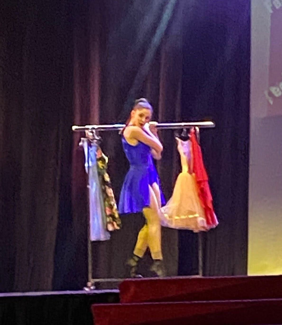 Una ballerina con un abito blu si trova dietro un appendiabiti su ruote che regge costumi su un palcoscenico con uno sfondo scuro.