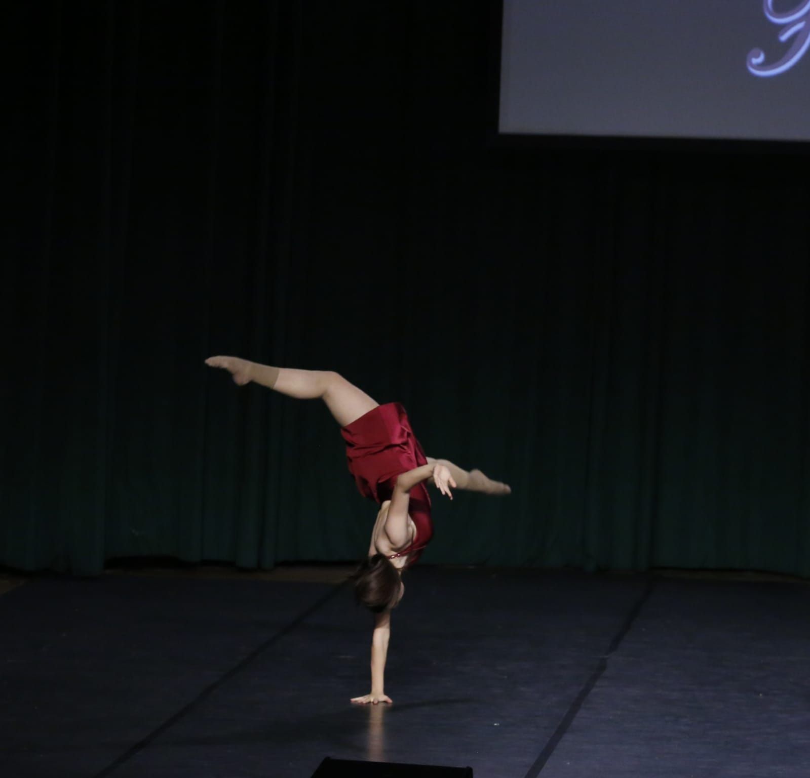 Su un palcoscenico buio, una ballerina con indosso un abito color bordeaux esegue una verticale su una mano, con le gambe distese.