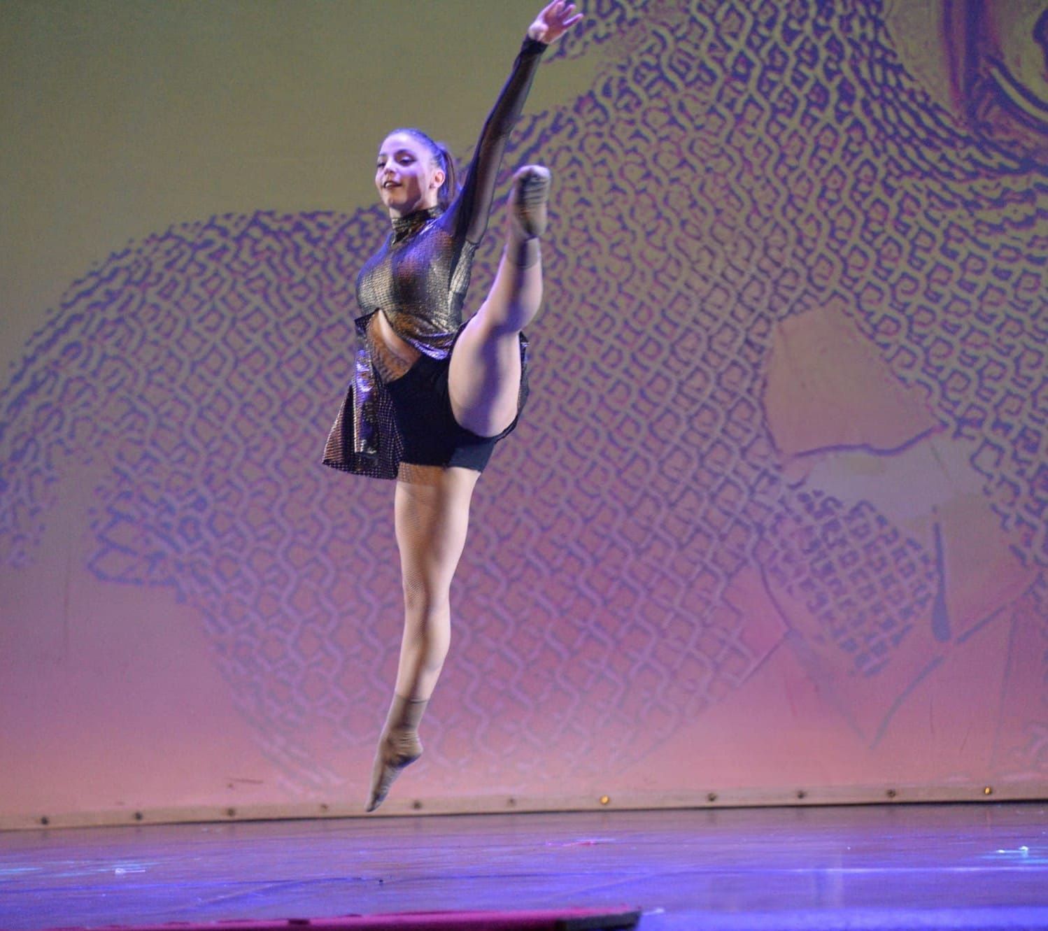 Una ballerina esegue un salto in spaccata laterale su uno sfondo decorato, in un palcoscenico scarsamente illuminato.