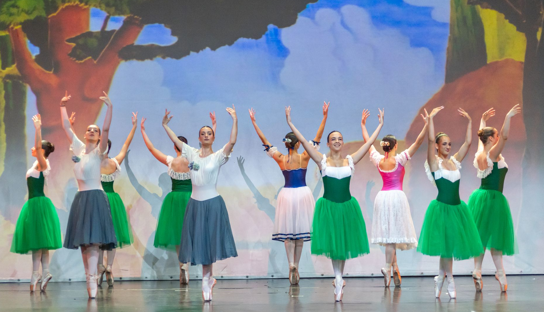 Un gruppo di ballerini di danza classica, con indosso top bianchi e gonne verdi o grigie, si esibisce sul palco con le braccia alzate, sullo sfondo di un dipinto.