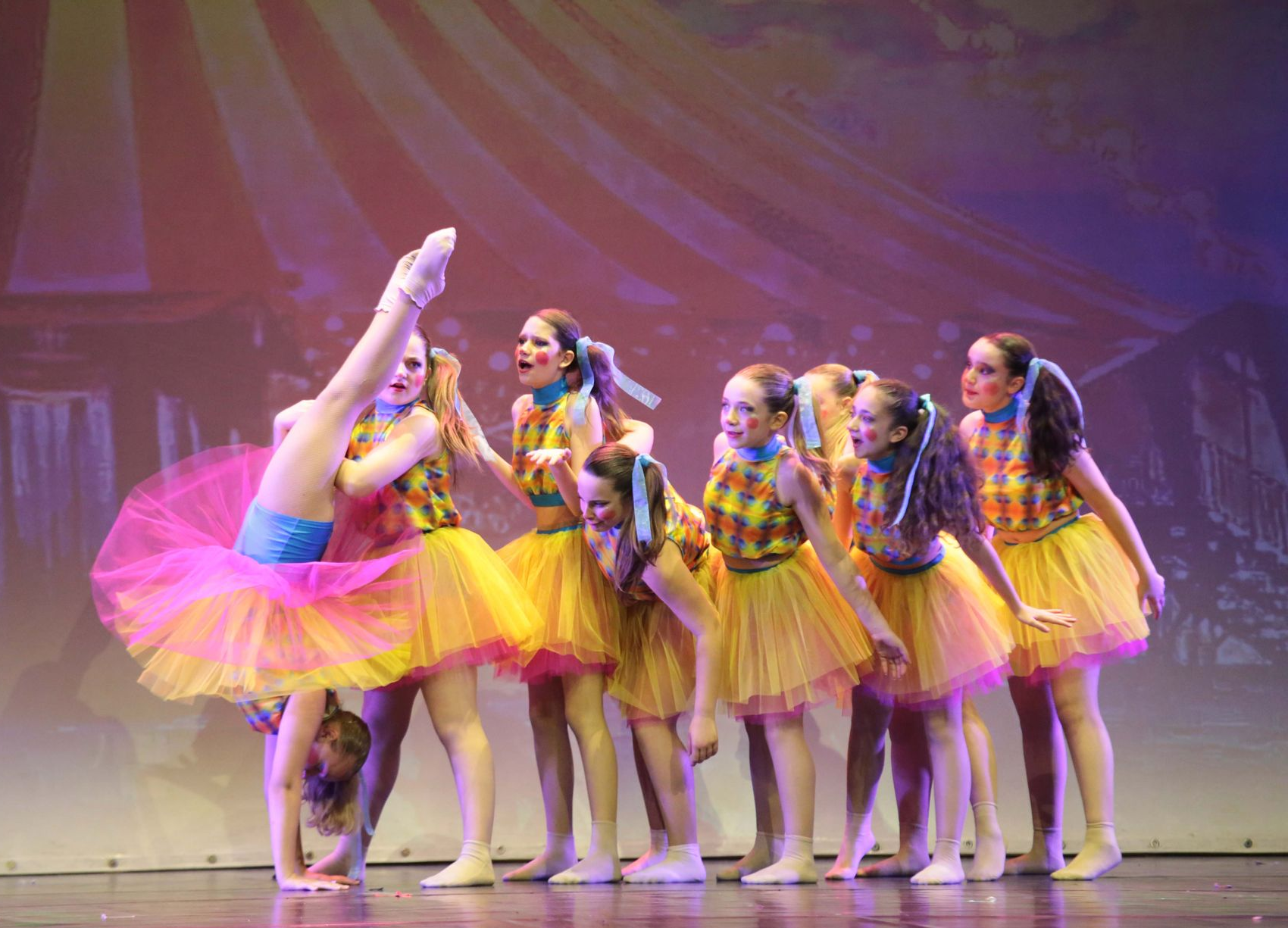 Ballerine in tutù gialli e rosa si esibiscono sul palco davanti a uno sfondo a tema circense, e una di loro esegue una verticale.
