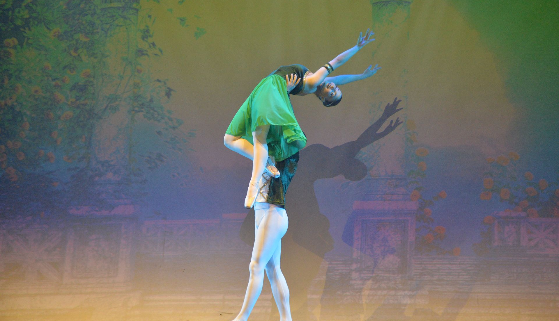 Una ballerina con un abito verde viene sollevata in alto dal suo partner, che indossa calzamaglie bianche, sullo sfondo di un palcoscenico nebuloso e suggestivo.
