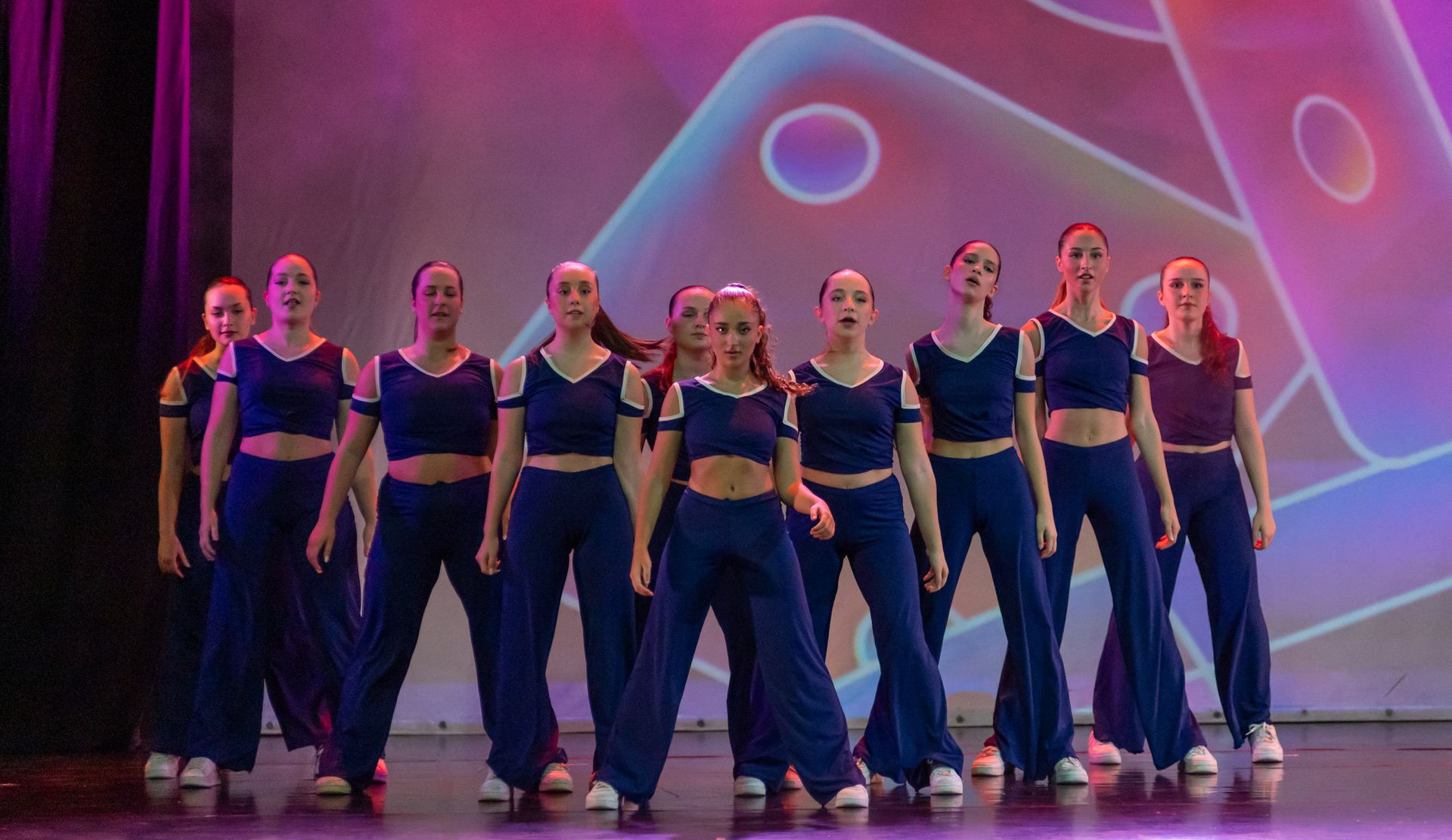 Un gruppo di ballerini, con indosso top corti blu scuro e pantaloni a gamba larga abbinati, si esibisce su un palco illuminato da una luce viola.