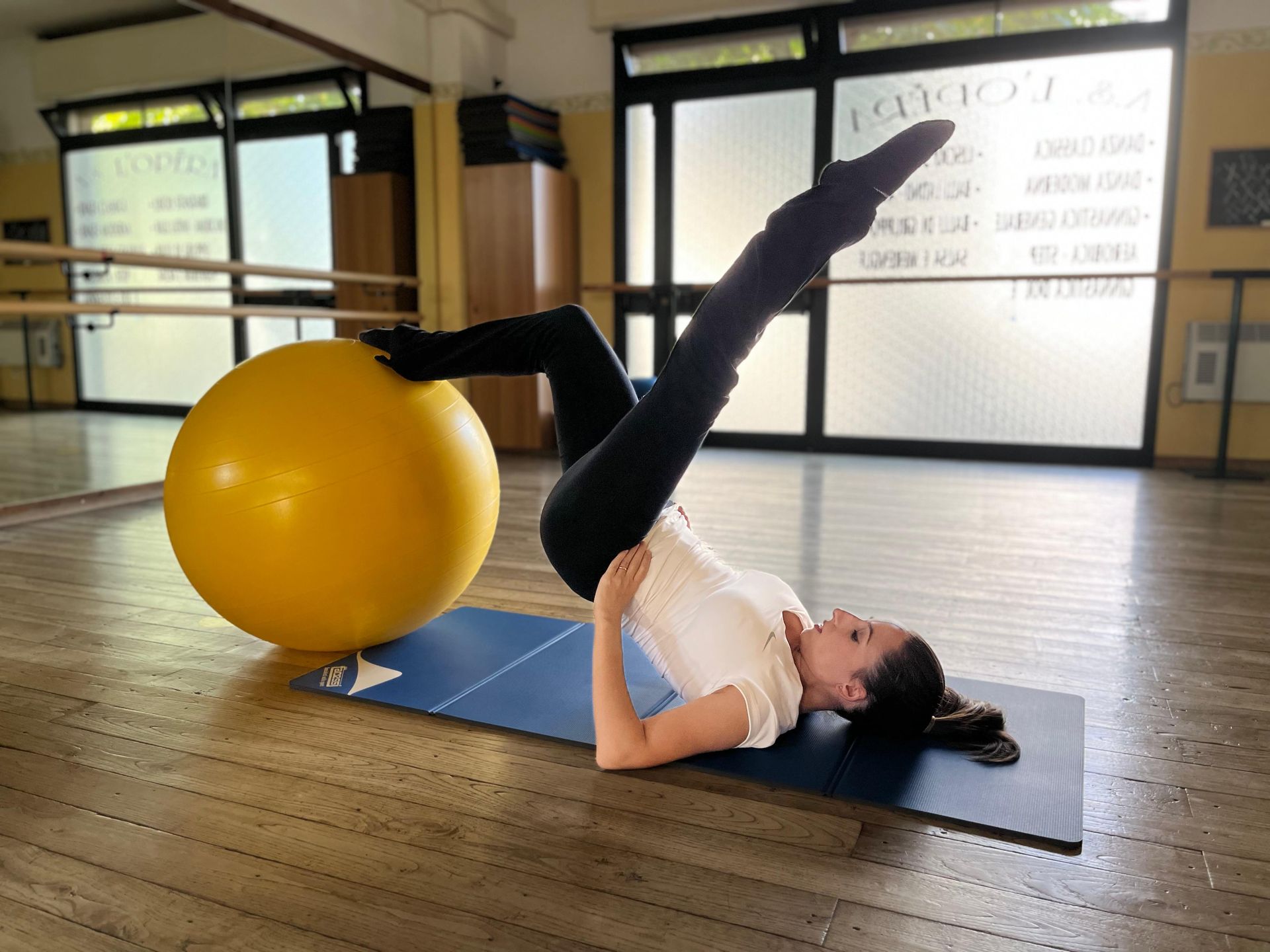 Una persona in una sala da ballo è sdraiata su un tappetino, in equilibrio sui gomiti con una gamba su una palla da ginnastica gialla e l'altra sollevata.