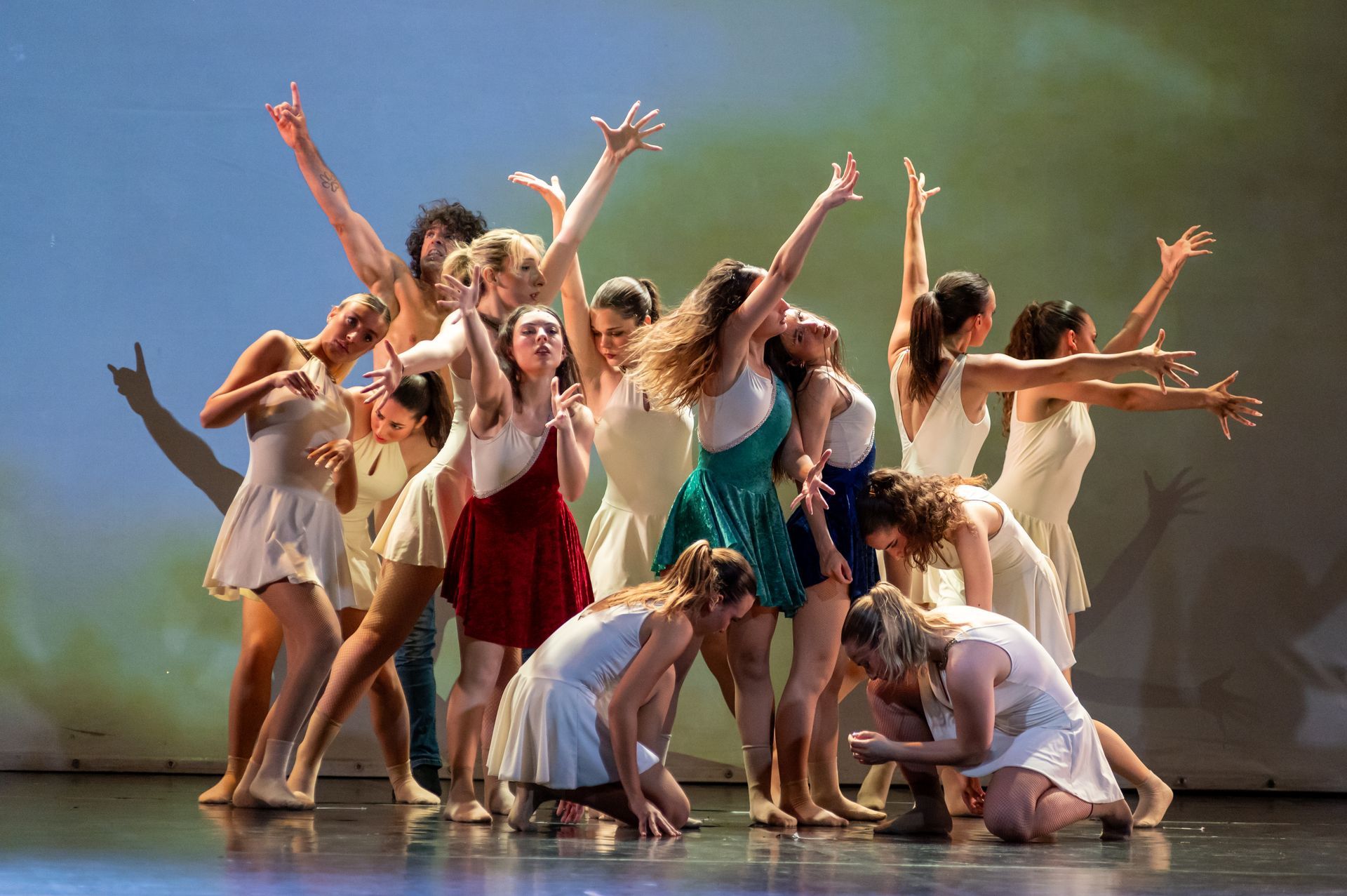 Un gruppo di ballerine in abiti femminili si esibisce su un palco con uno sfondo astratto, posando con le braccia alzate all'unisono.