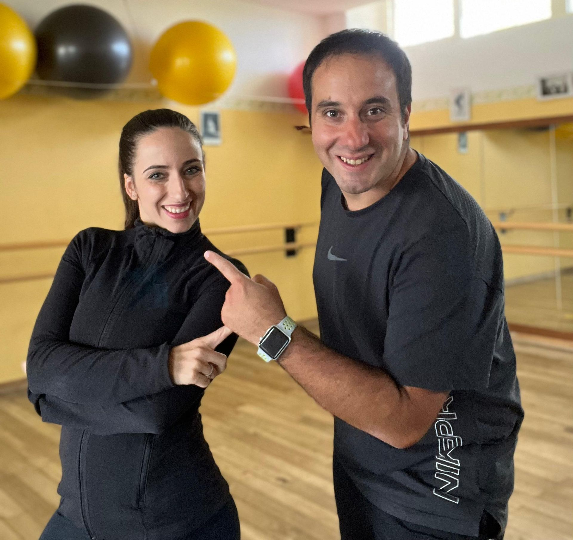 Una donna e un uomo sorridenti in una palestra. L'uomo indica il braccio della donna; entrambi indossano abbigliamento sportivo.