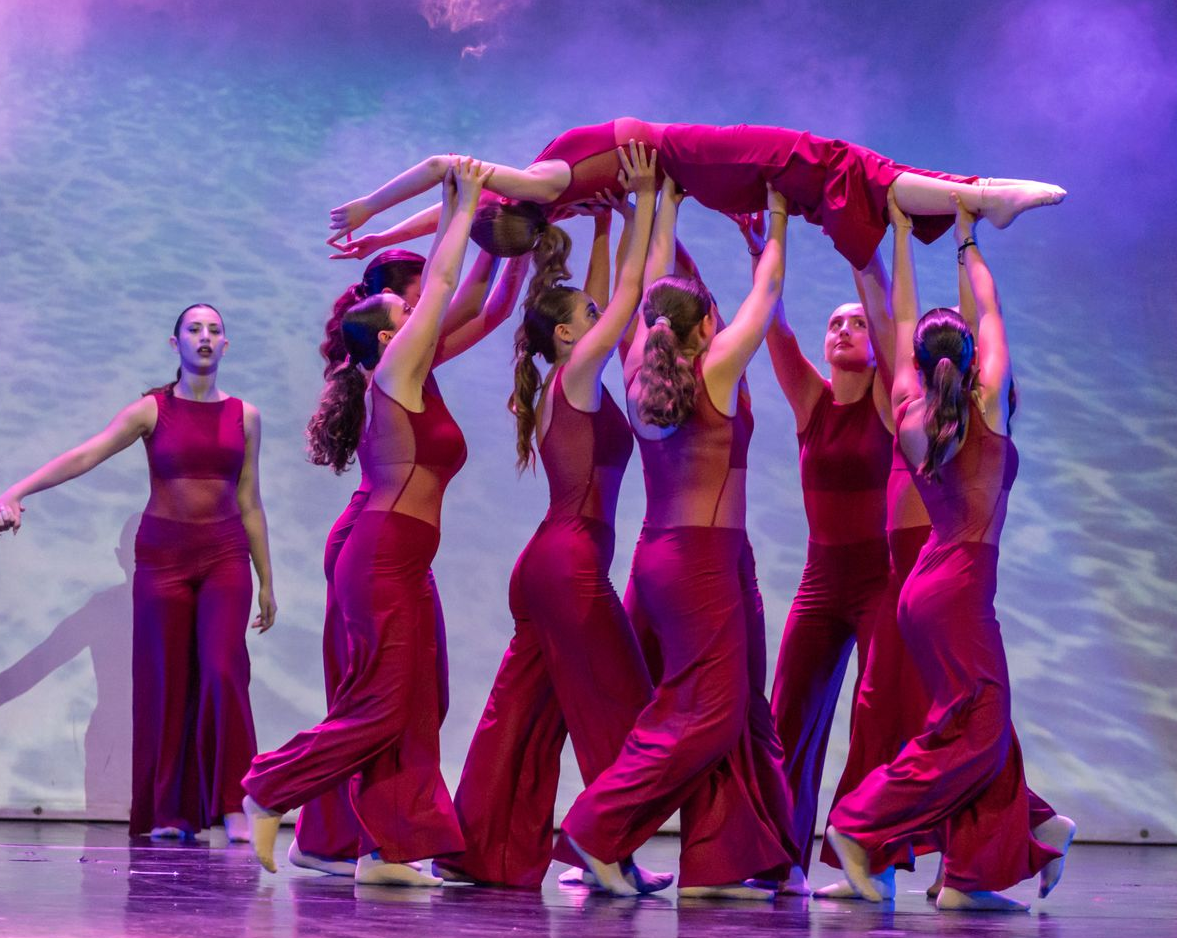Un gruppo di ballerini con indosso abiti magenta coordinati esegue un sollevamento, con un ballerino tenuto orizzontalmente sopra gli altri.