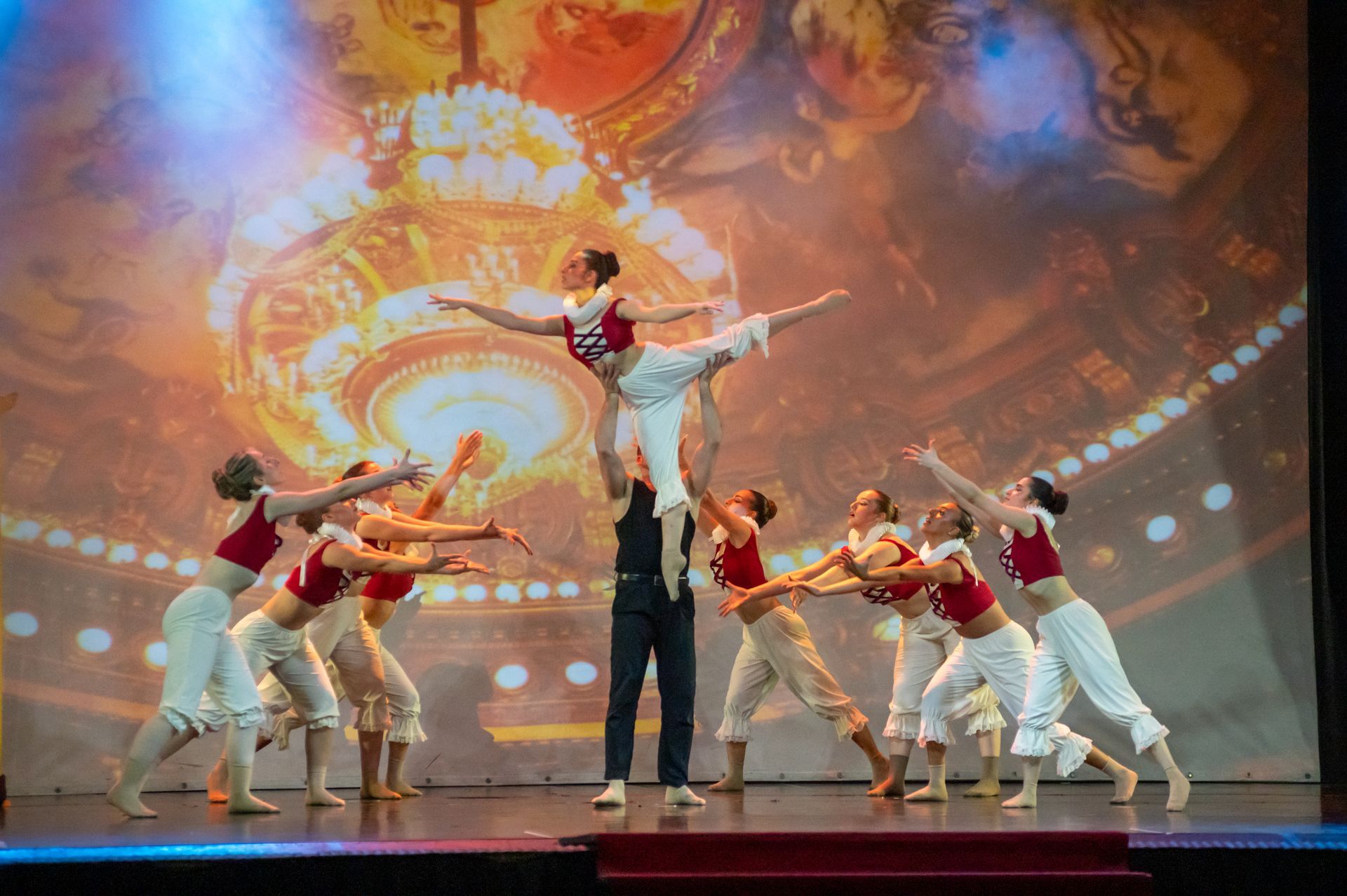 Un gruppo di ballerini sul palco, con un ballerino che solleva una ballerina sopra la sua testa davanti a uno sfondo dorato.