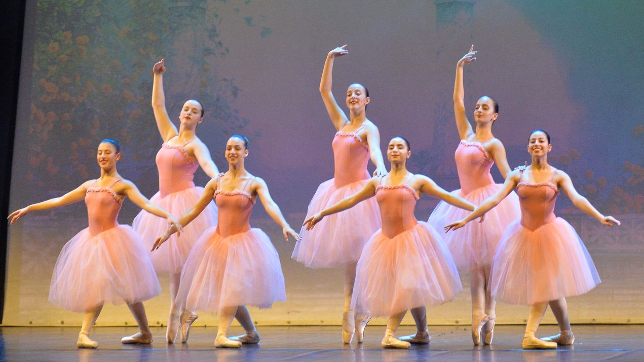 Sette ballerine in tutù rosa posano su un palcoscenico, con le braccia tese in varie posizioni aggraziate.