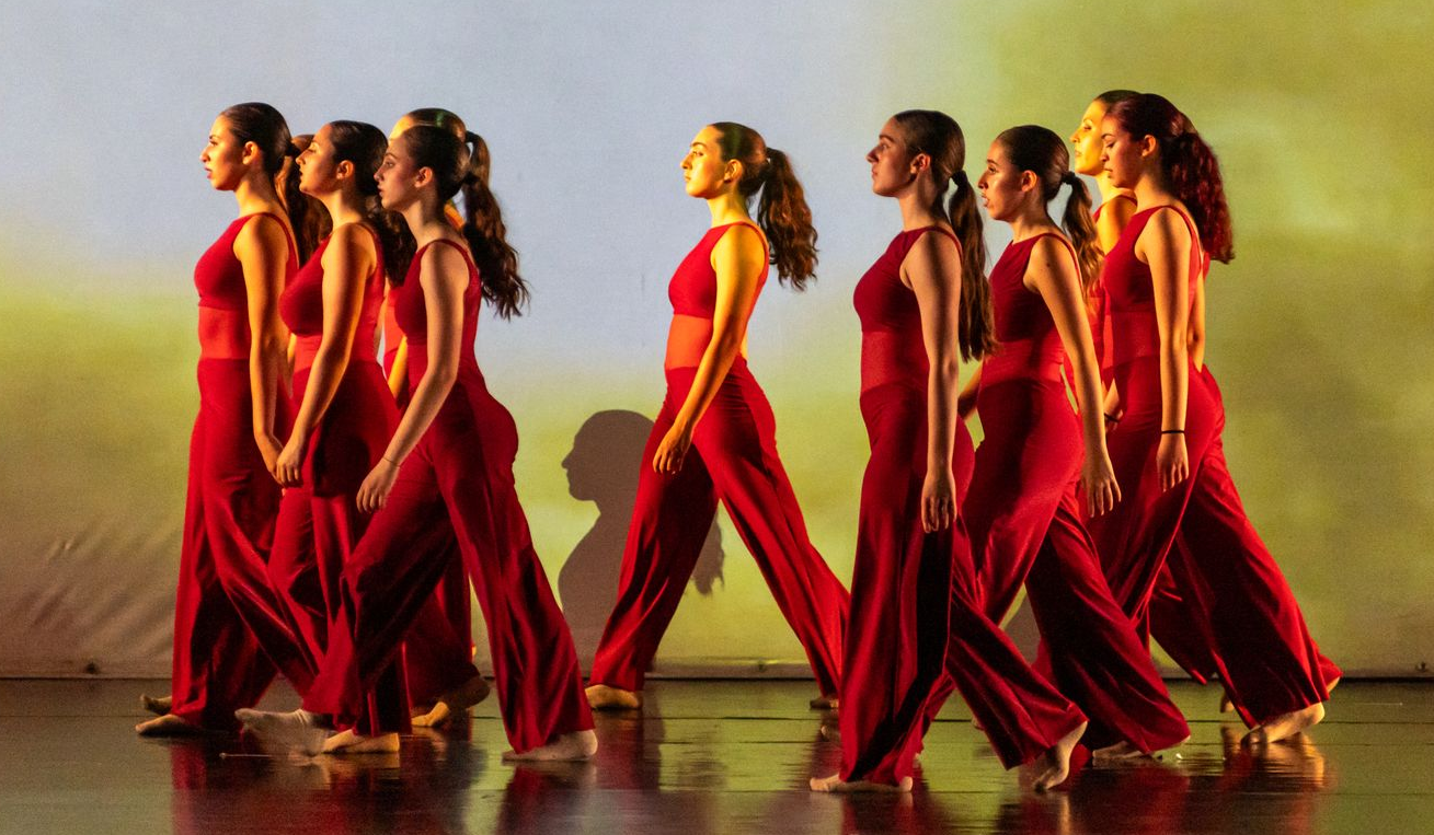 Una fila di artisti in abiti rossi attraversa un palco contro uno sfondo giallo e sfocato.