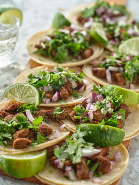 Tacos with cilantro, onions, and lime wedges on a wooden board.