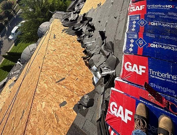 Roof under repair with exposed plywood, black shingles, and stacked GAF Timberline shingles