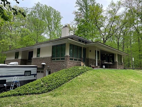 Suburban brick ranch house with green trim and covered porch on a grassy hill surrounded by trees