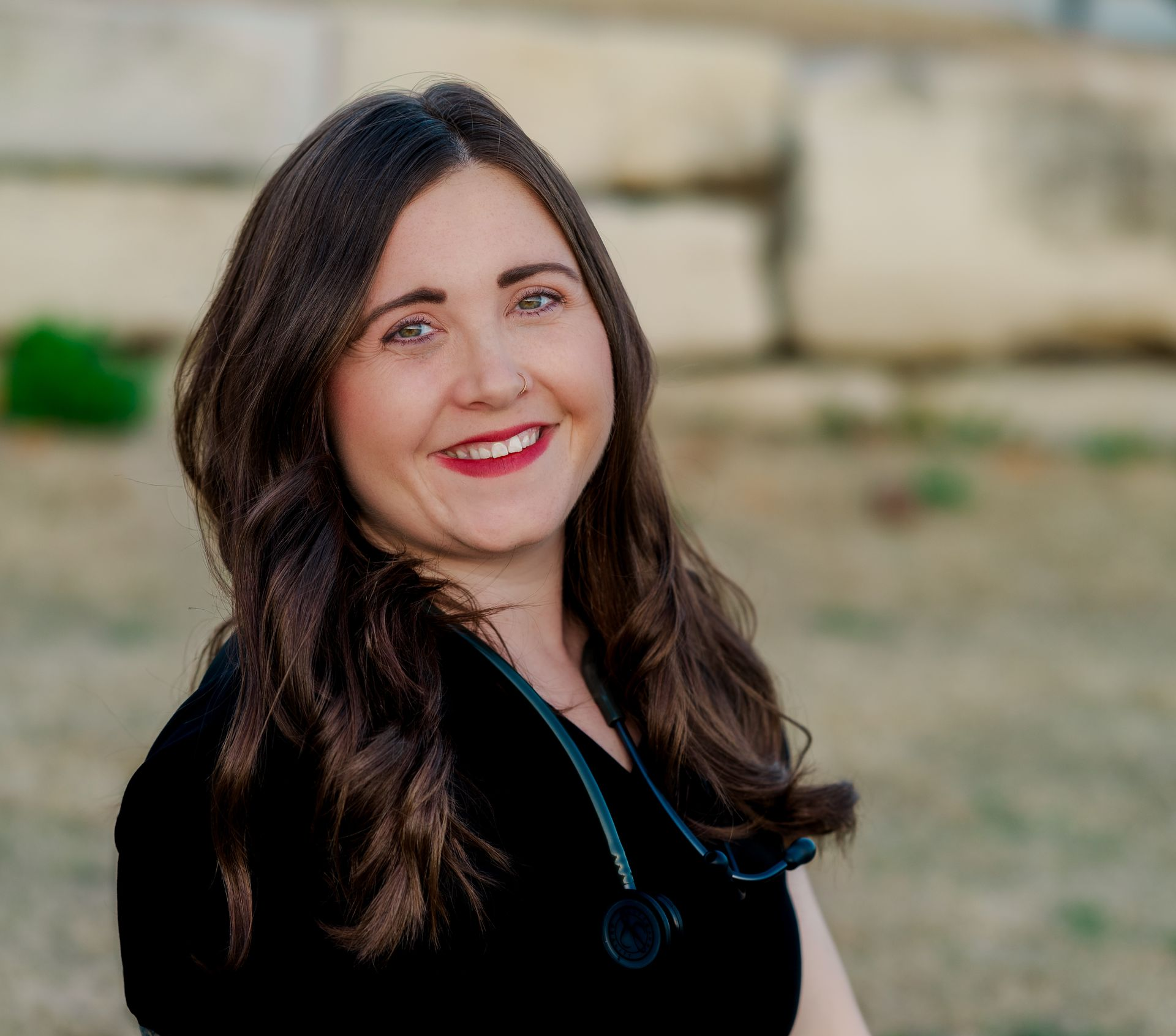A smiling medical professional with long brown hair, wearing black scrubs and a stethoscope, posing outdoors.