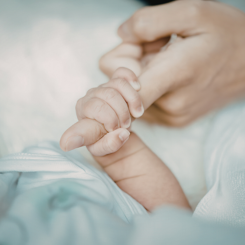 A small baby's hand grasping an adult's finger against a soft, light blue background.