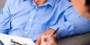 a man in a blue shirt is sitting on a couch while a woman writes on a piece of paper .