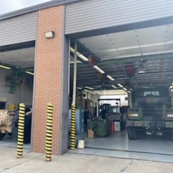 Exterior of a vehicle maintenance bay. Vehicles are inside, with open garage doors. Yellow and black poles stand in front.