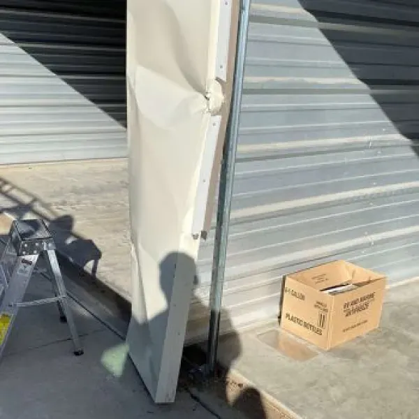Damaged storage unit door with crumpled beige panel, metal frame, and cardboard box on concrete.