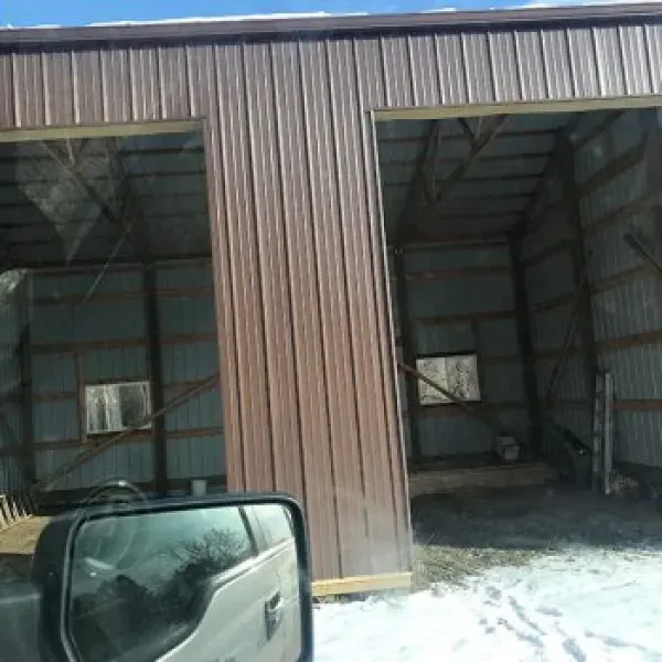 Brown metal building with two open bays, snow on the ground, and a vehicle's side mirror in view.