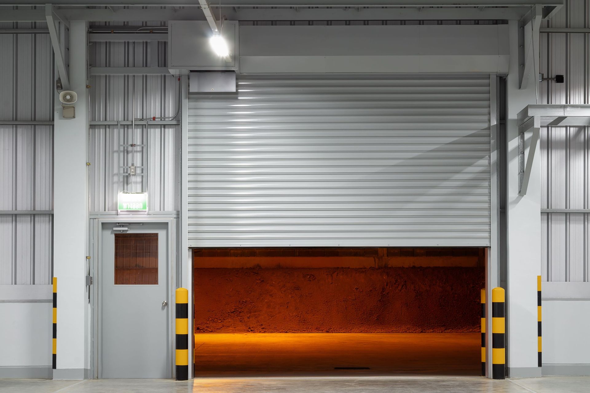 Roller shutter partially open, revealing an interior with warm lighting. Door and yellow/black bollards on sides.