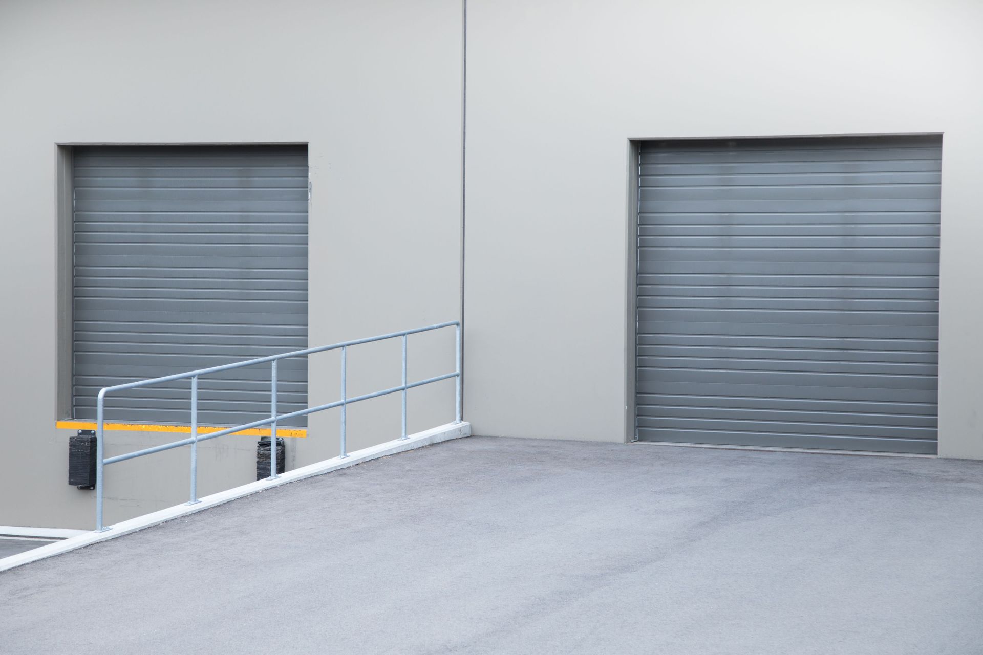Gray industrial building with two closed roll-up doors, a ramp, and metal railing.