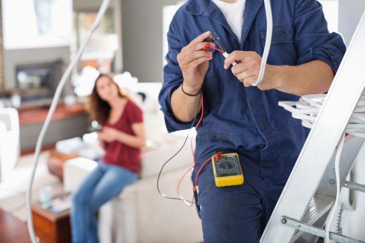 An electrician is working on a wire in a living room while a woman sits on a couch.