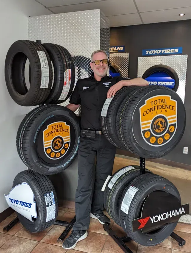 Un homme en uniforme noir, souriant, pose devant un présentoir de pneus dans un magasin automobile.