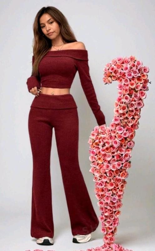 Una mujer con un top y pantalones color granate con hombros descubiertos posa junto a una escultura alta cubierta de flores.