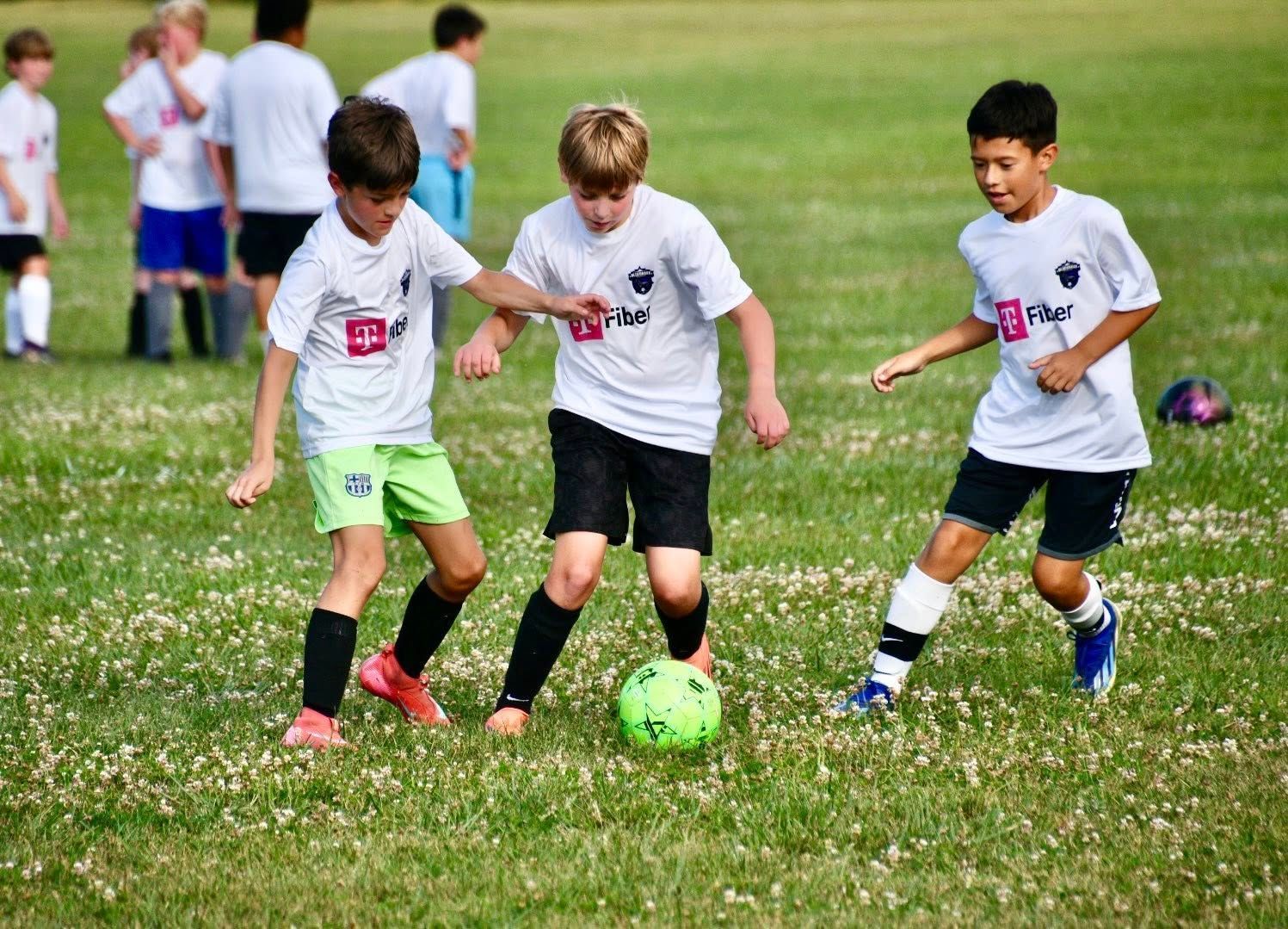 Three young boys playing soccer on a grassy field; one dribbles the ball, two others try to take it.