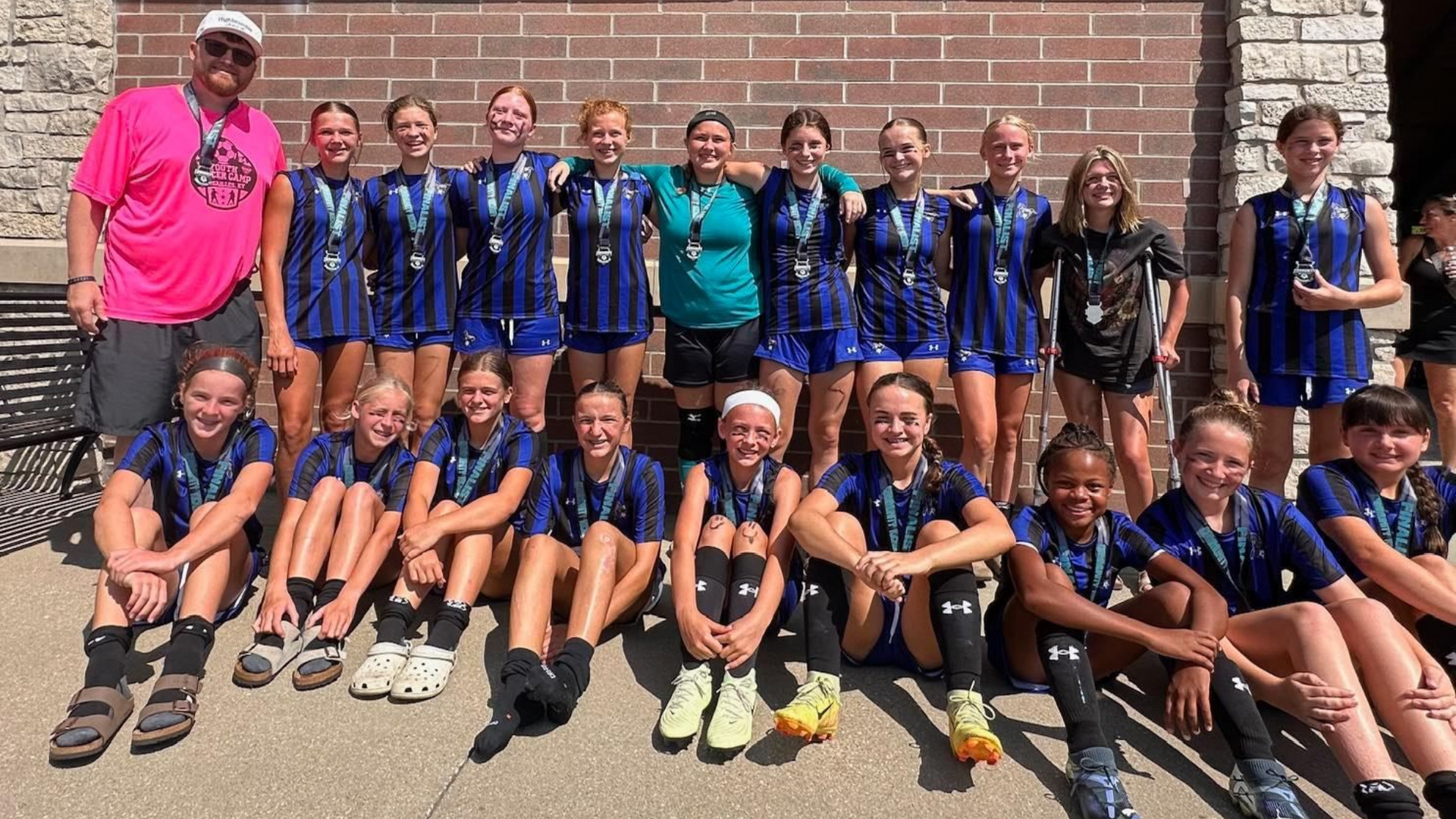 Youth girls volleyball team posing for a group photo with medals. Brick wall, sunny outdoors.