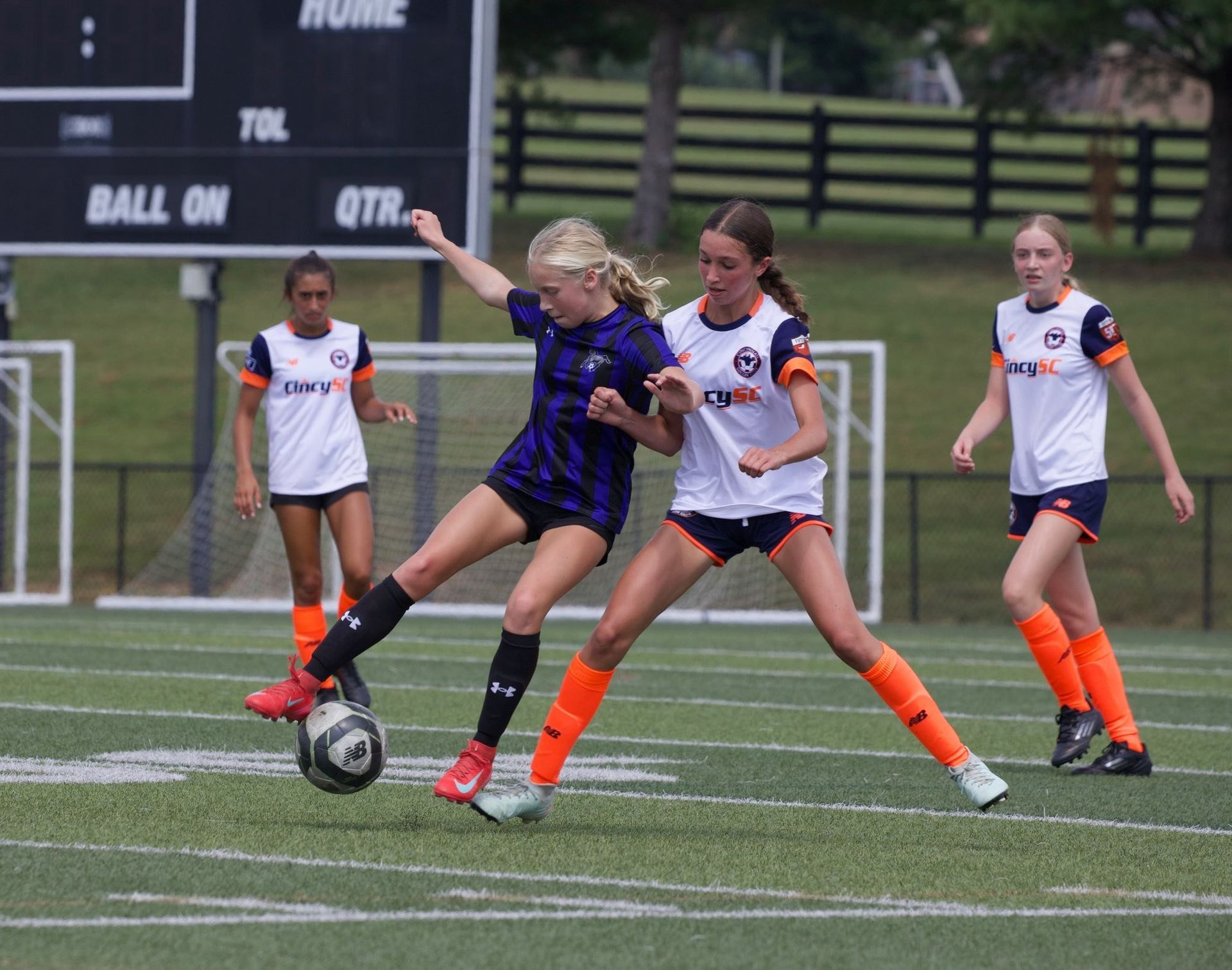 Soccer players in action on a field. One in purple kicks the ball, two in white attempt to defend.