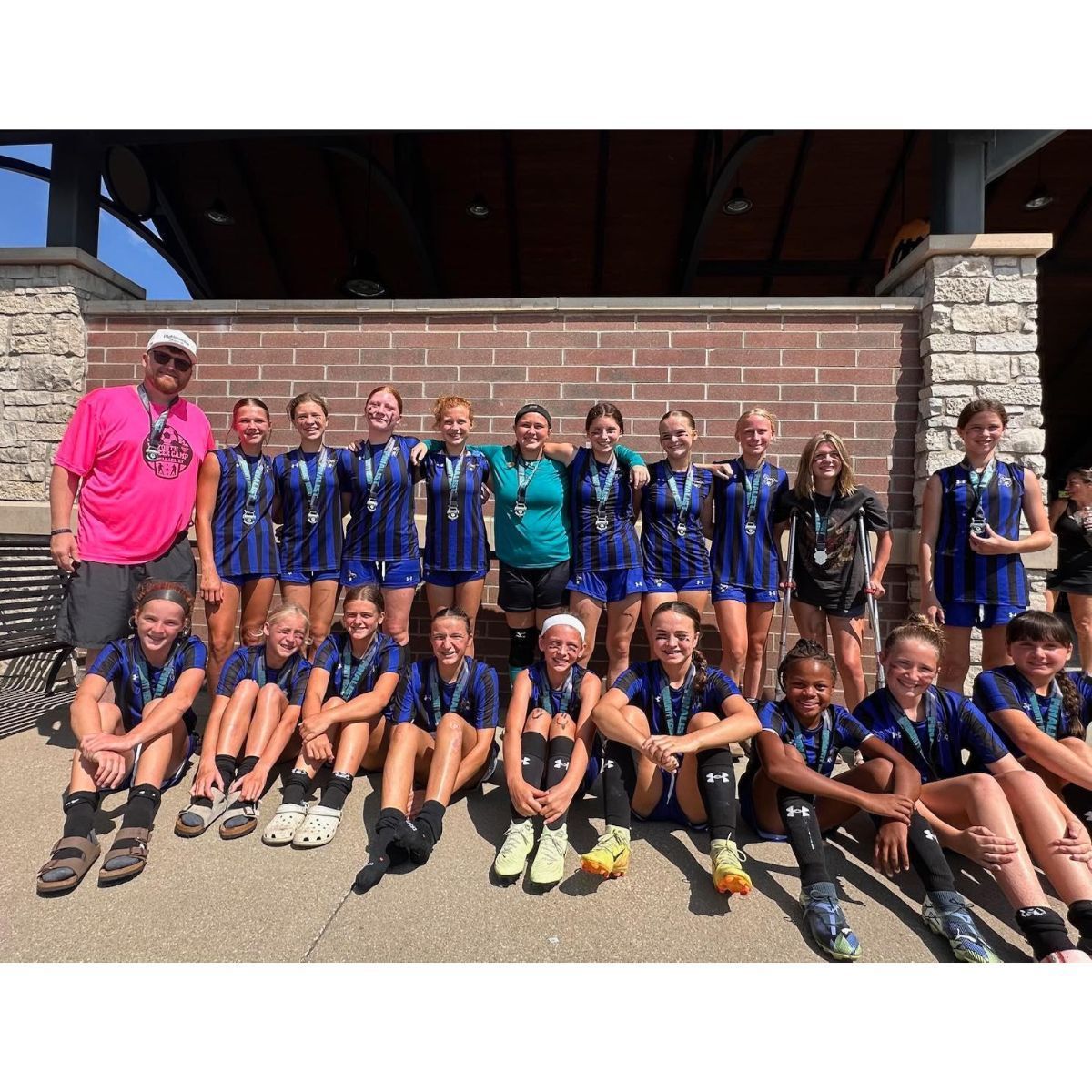 Youth girls volleyball team posing for a group photo with medals. Brick wall, sunny outdoors.