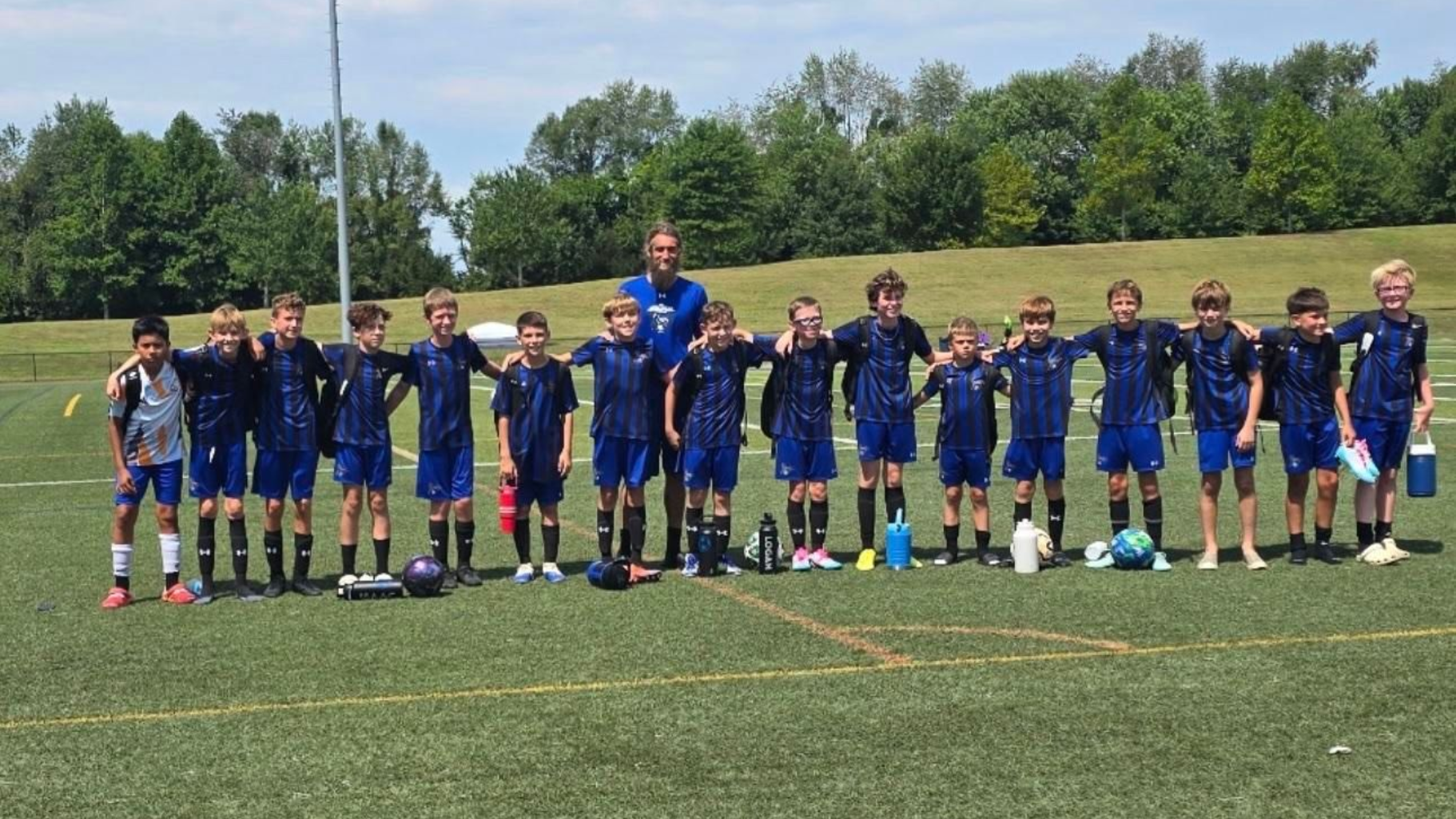Youth soccer team posing on a field; blue uniforms, two adults, green grass, cloudy sky.