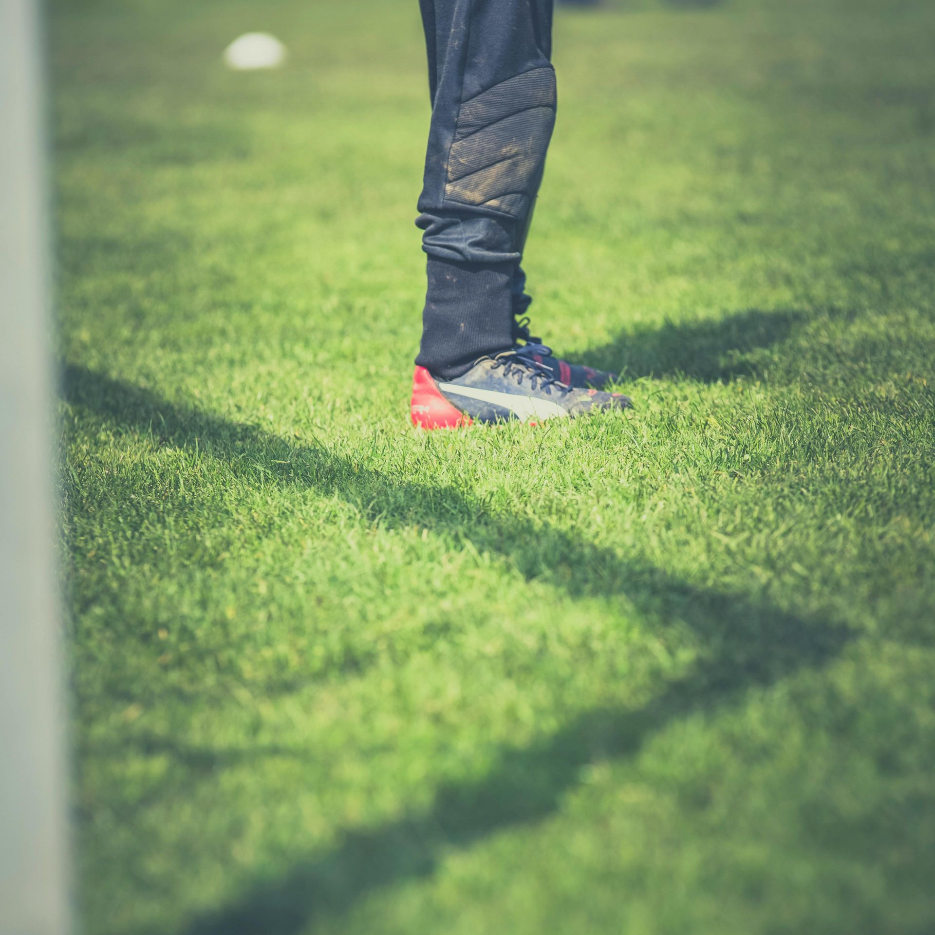 Soccer player's foot in cleats, standing on green grass field. Black pants, red and black shoe, long shadow.