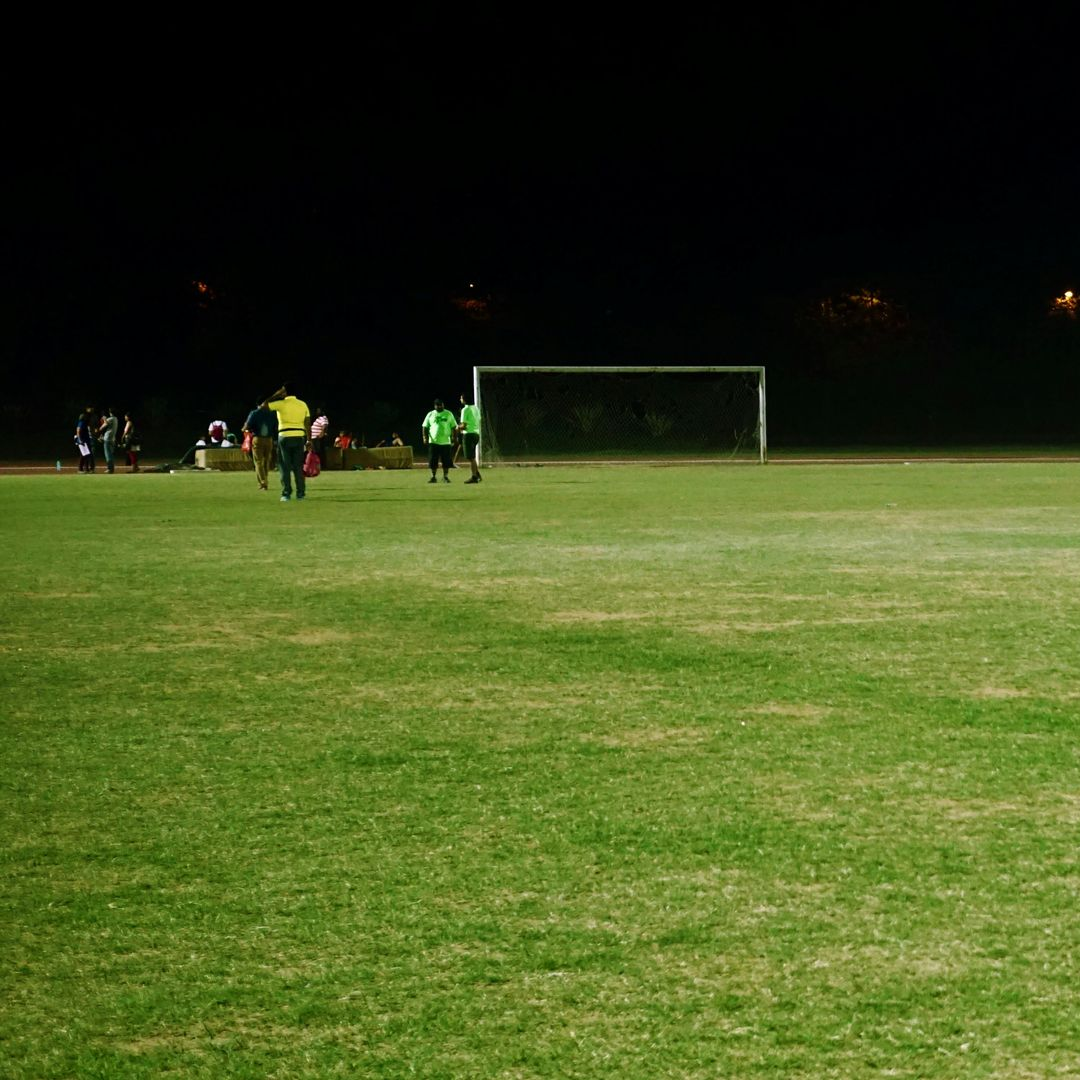 Soccer game at night: Green field, players near goal. Dark sky, illuminated by stadium lights.