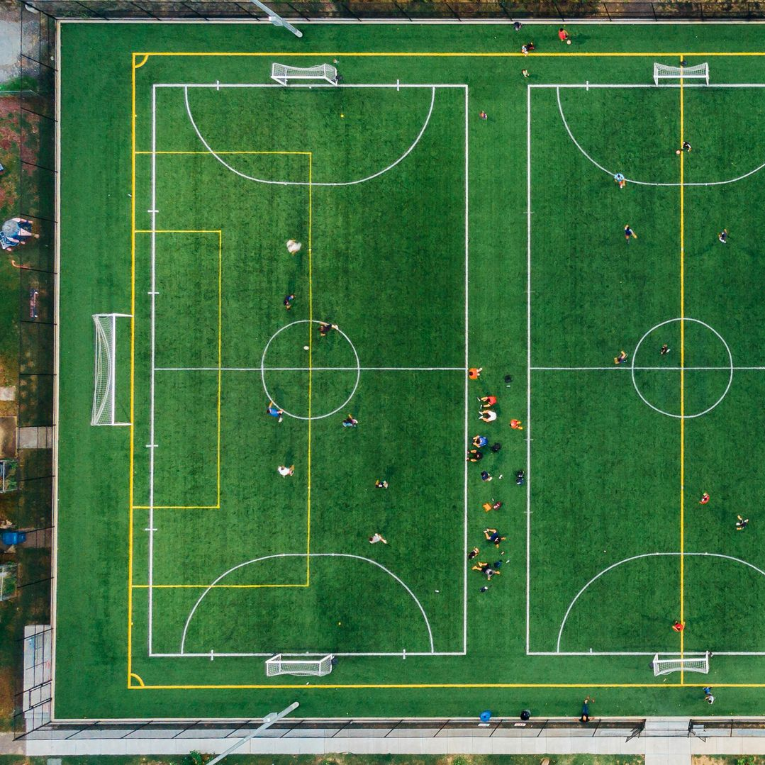 Aerial view of two green soccer fields with players, outlined with white and yellow lines.