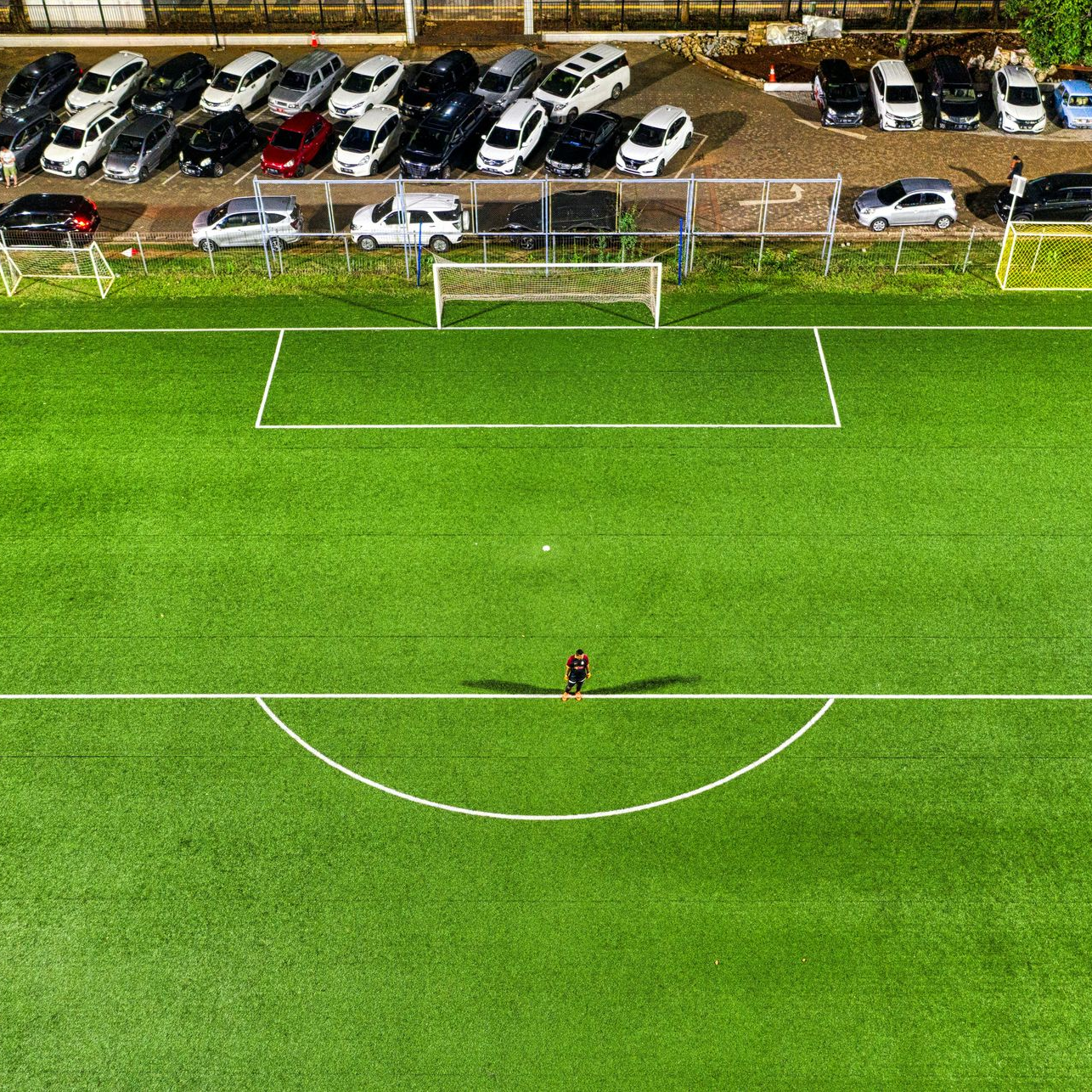 Soccer field with a lone player, cars parked nearby, and a green field.