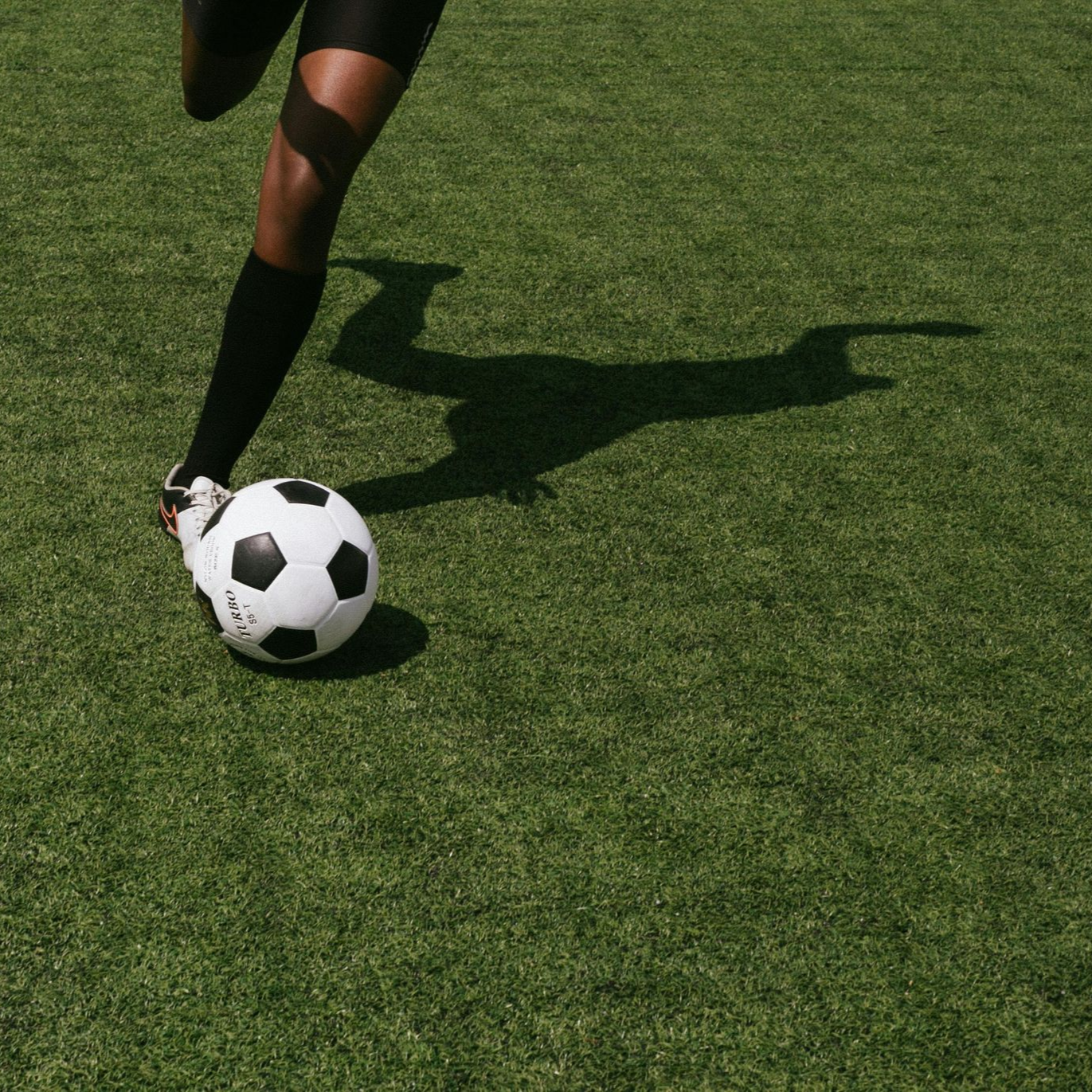 Soccer player's leg kicking a soccer ball on a green field, with a long shadow.