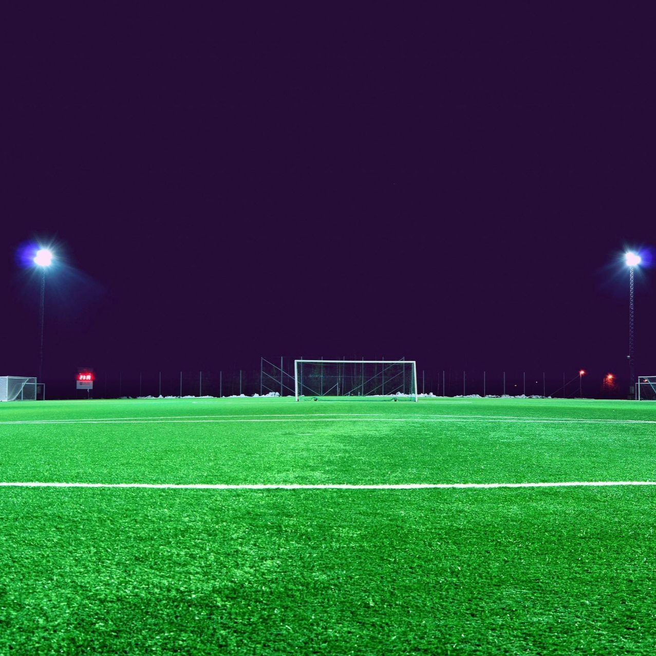 Soccer field at night under bright lights, with goal posts and a dark purple sky.