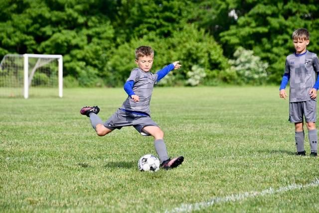 Boy kicking a soccer ball on a green field, another boy watches, goal in background.