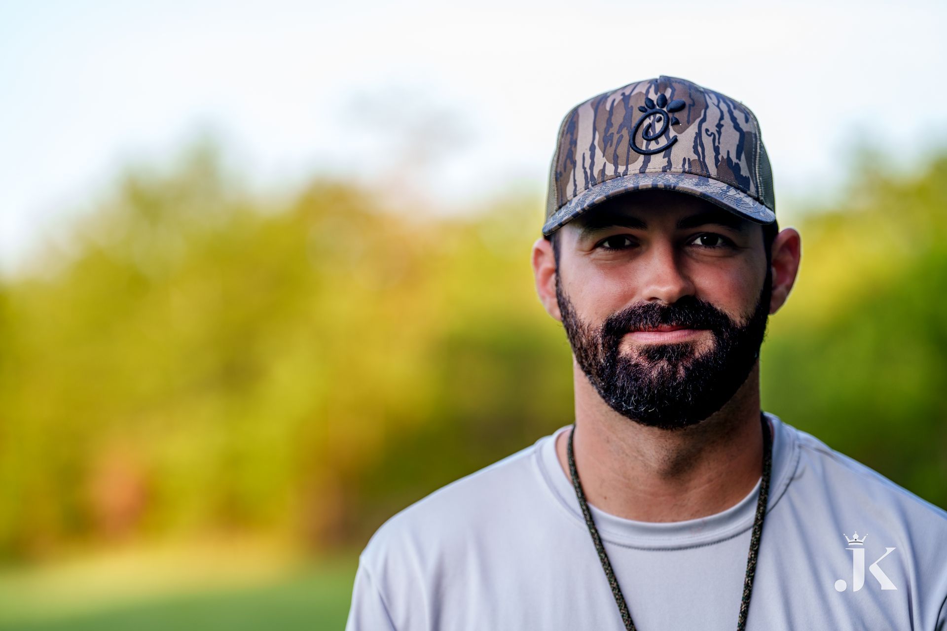 Man with beard wearing camo hat and grey shirt, outdoors with blurred background.