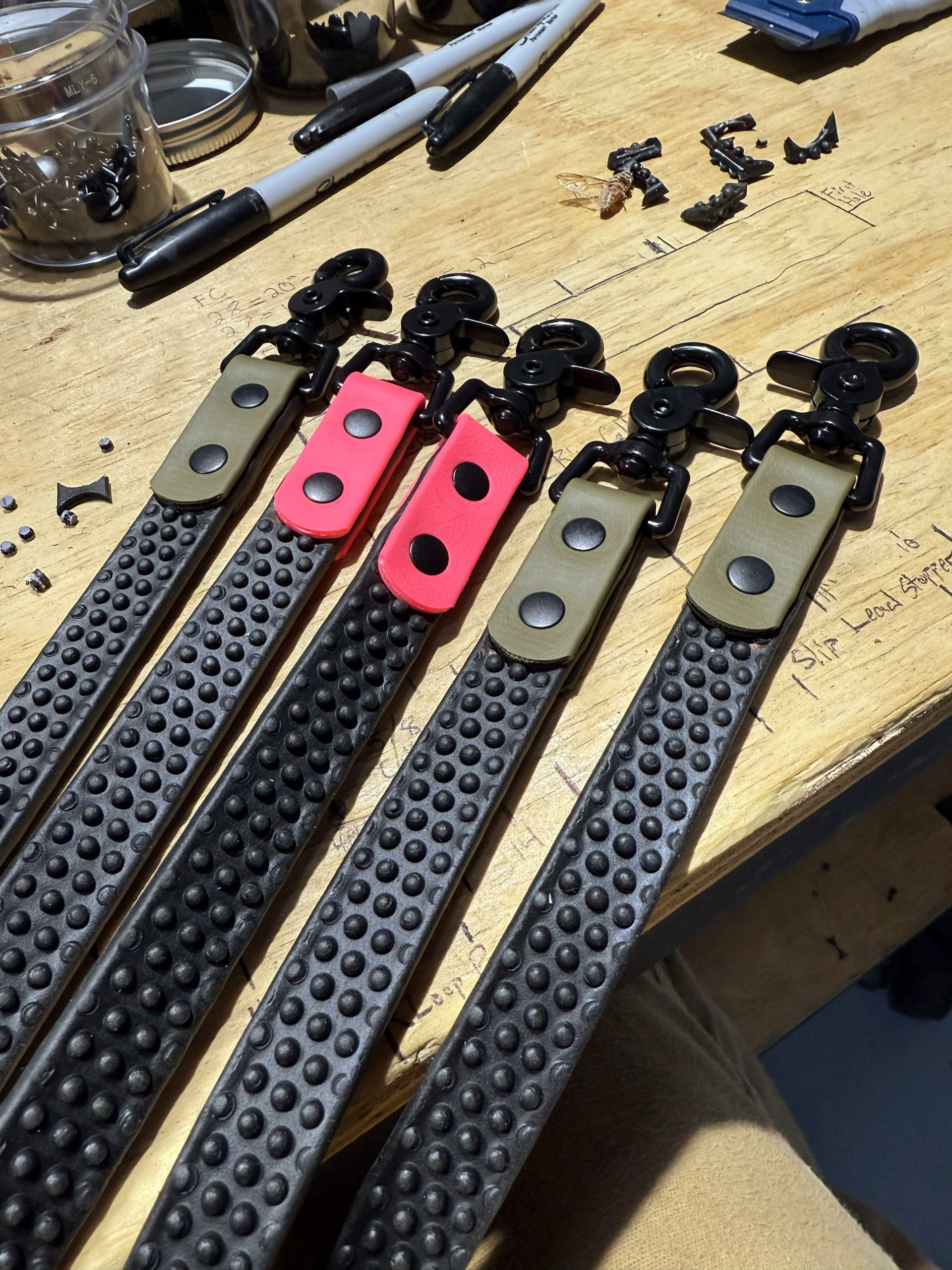 Five black studded straps with varying colored buckles and black carabiners on a wooden surface.