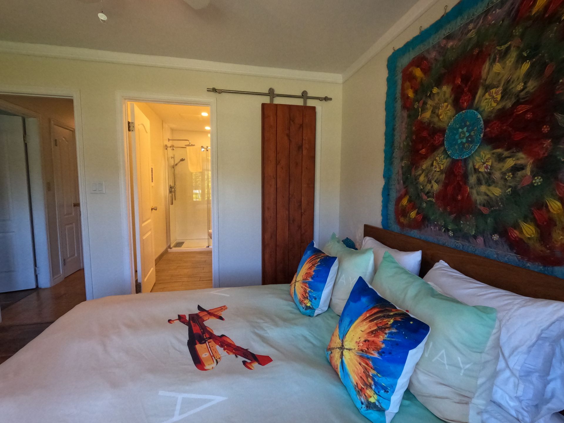 Bright bedroom with a large bed, colorful pillows, and abstract wall art near an open doorway.