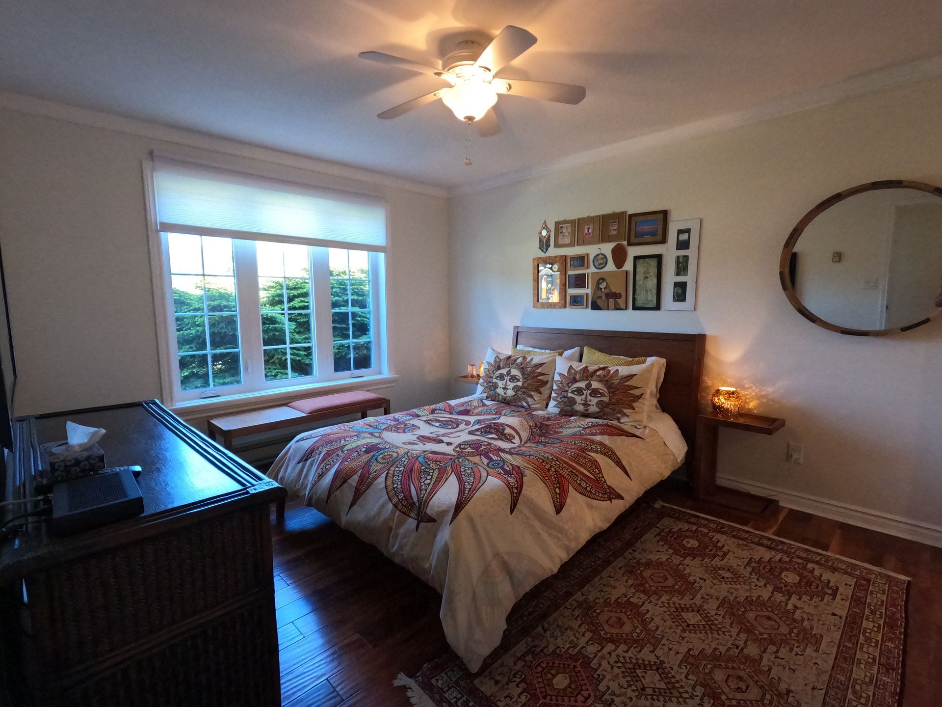 Cozy bedroom with a made bed, large window, dresser, rug, and wall mirror