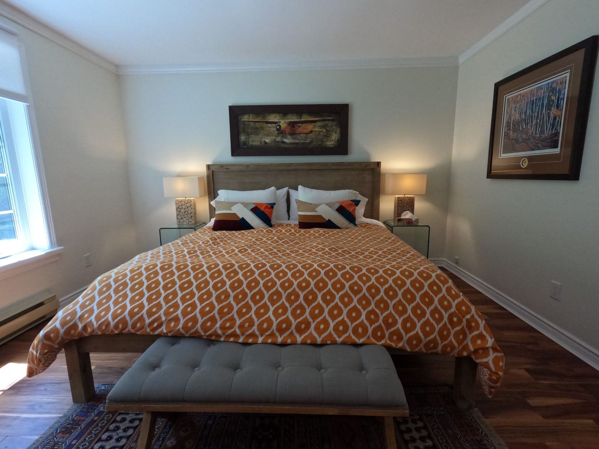 Neatly made bedroom with a bed, orange patterned comforter, bedside lamps, and framed wall art.